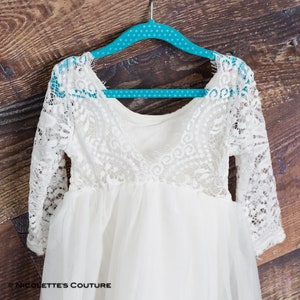 Puede incluir: Vestido de beb&eacute; blanco de encaje y tul con mangas largas colgado de una percha de lunares turquesa. El vestido tiene un escote redondo y una falda fruncida.