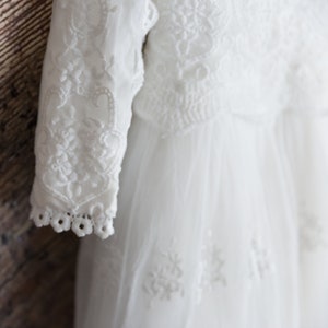 May include: White lace baby dress with long sleeves and a delicate floral pattern. The dress is made of soft tulle and has a flowing skirt.