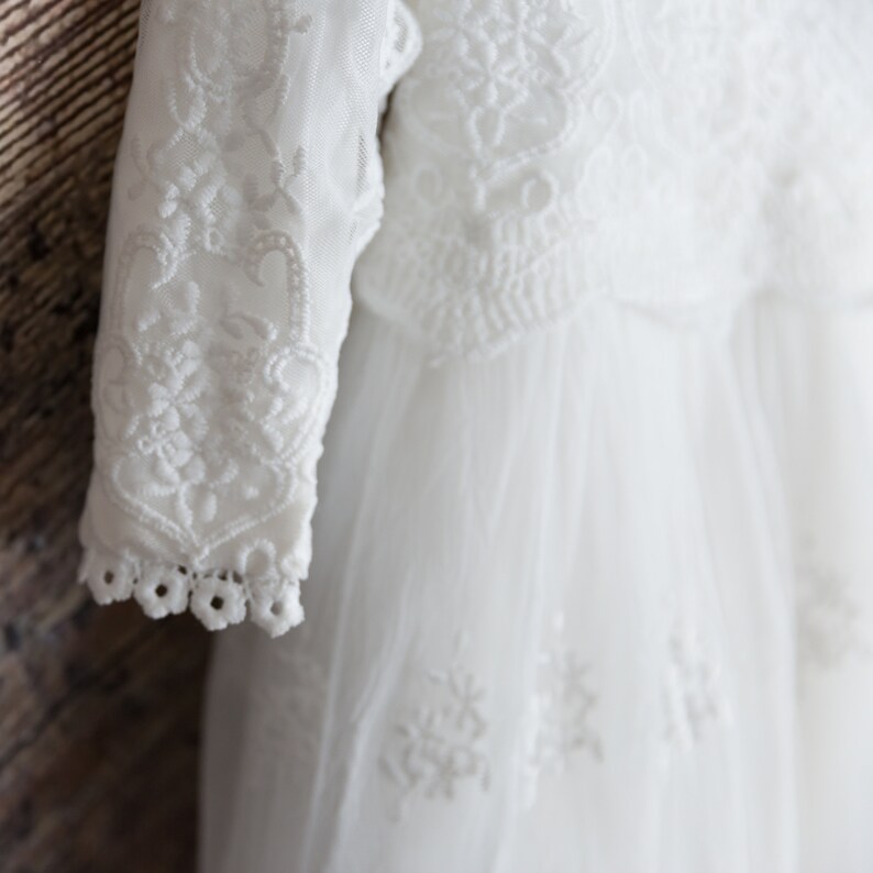 May include: White lace baby dress with long sleeves and a delicate floral pattern. The dress is made of soft tulle and has a flowing skirt.
