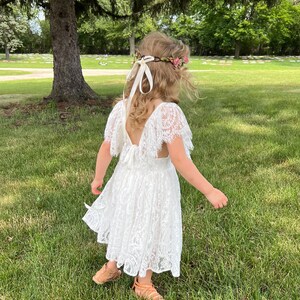 May include: A young girl wearing a white lace dress with a floral crown and a white ribbon. She is standing in a grassy field with trees in the background.