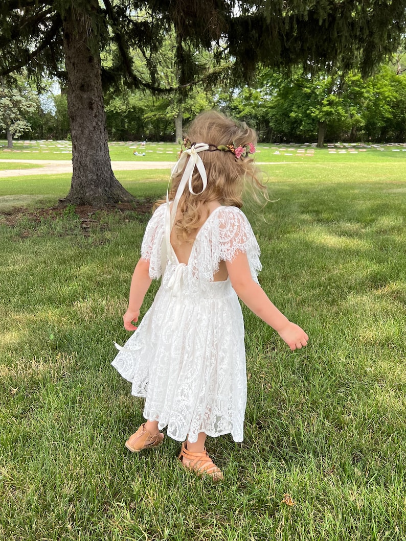 May include: A young girl wearing a white lace dress with a floral crown and a white ribbon. She is standing in a grassy field with trees in the background.