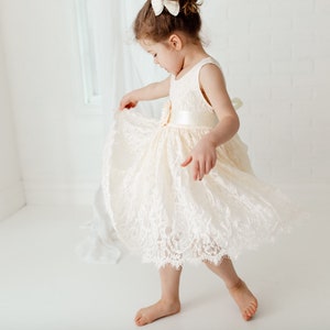 May include: A young girl wearing a white lace dress with a satin sash and a bow in her hair. She is twirling and smiling.