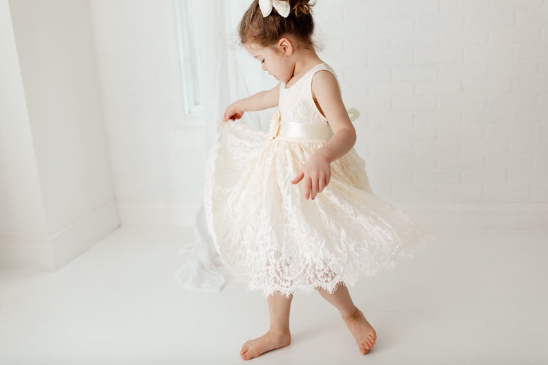 May include: A young girl wearing a white lace dress with a satin sash and a bow in her hair. She is twirling and smiling.