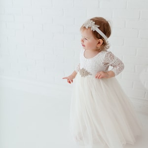 Pu&ograve; includere: Una bambina che indossa un abito bianco in pizzo con gonna di tulle e un cerchietto scintillante. L'abito ha un corpetto in pizzo e una cintura in raso con una fibbia gioiello.