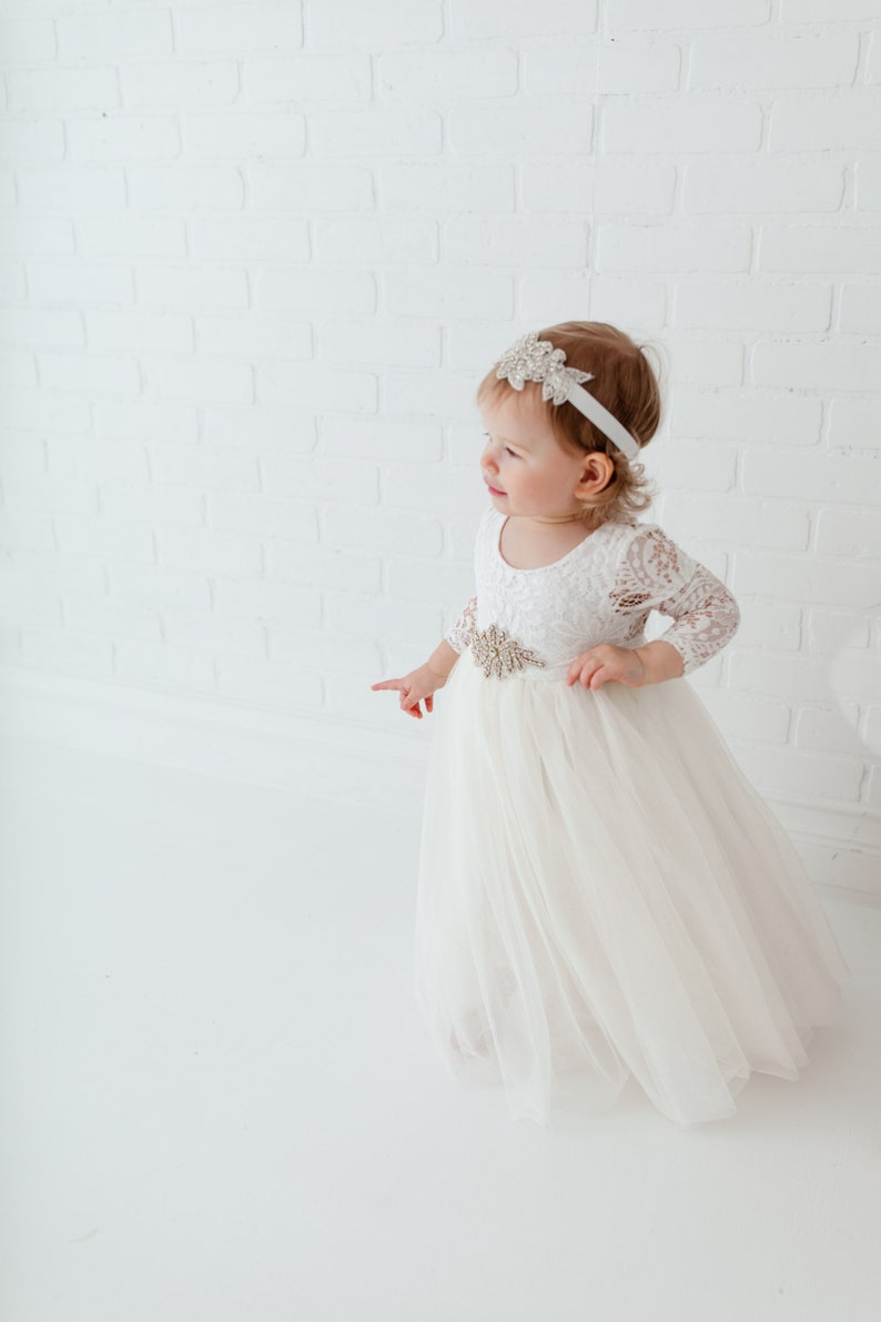 Pu&ograve; includere: Una bambina che indossa un abito bianco in pizzo con gonna di tulle e un cerchietto scintillante. L'abito ha un corpetto in pizzo e una cintura in raso con una fibbia gioiello.