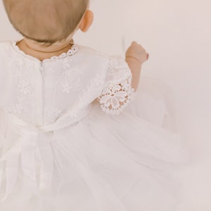 K&ouml;nnte beinhalten: Ein wei&szlig;es Babykleid mit Spitzenbesatz und einer Schleife aus Satin. Das Kleid ist mit kleinen Blumenmustern bestickt.