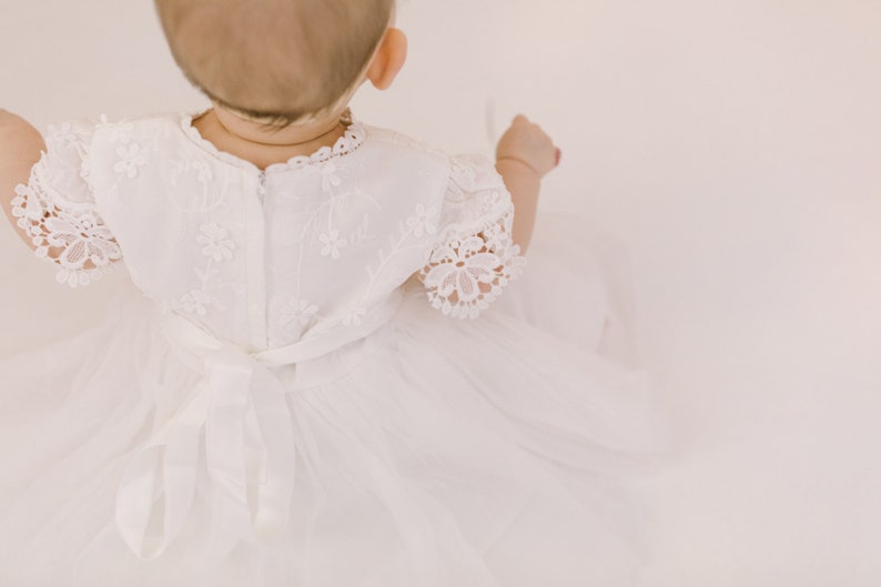 K&ouml;nnte beinhalten: Ein wei&szlig;es Babykleid mit Spitzenbesatz und einer Schleife aus Satin. Das Kleid ist mit kleinen Blumenmustern bestickt.