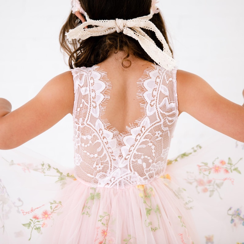 May include: A sleeveless dress featuring a white lace bodice with a deep V-shaped back and a light pink tulle skirt adorned with embroidered floral patterns. The garment is paired with a matching cream-coloured lace hair accessory tied with a bow.