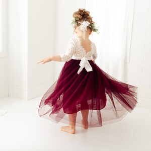May include: A young girl wearing a white lace top with a burgundy tulle skirt. The skirt is flowing and the girl is twirling. She is wearing a flower crown.