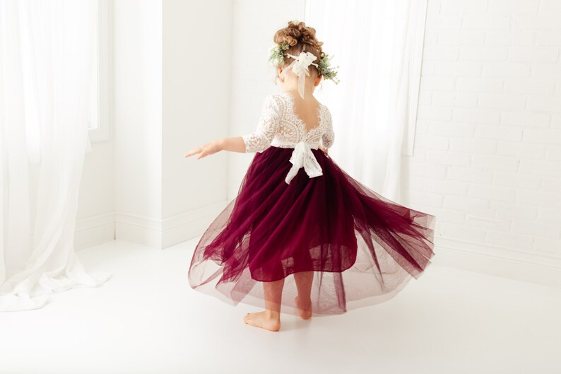 May include: A young girl wearing a white lace top with a burgundy tulle skirt. The skirt is flowing and the girl is twirling. She is wearing a flower crown.