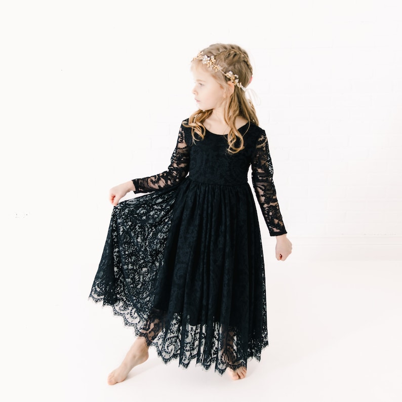 May include: A black long-sleeved dress featuring an intricate floral lace overlay with a scalloped hemline. The dress has a round neckline and a fitted bodice that transitions into a full, flowing skirt. A delicate floral headpiece is worn.