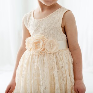 May include: A white lace dress with a peach-colored flower sash. The dress has a sleeveless design and a full skirt.
