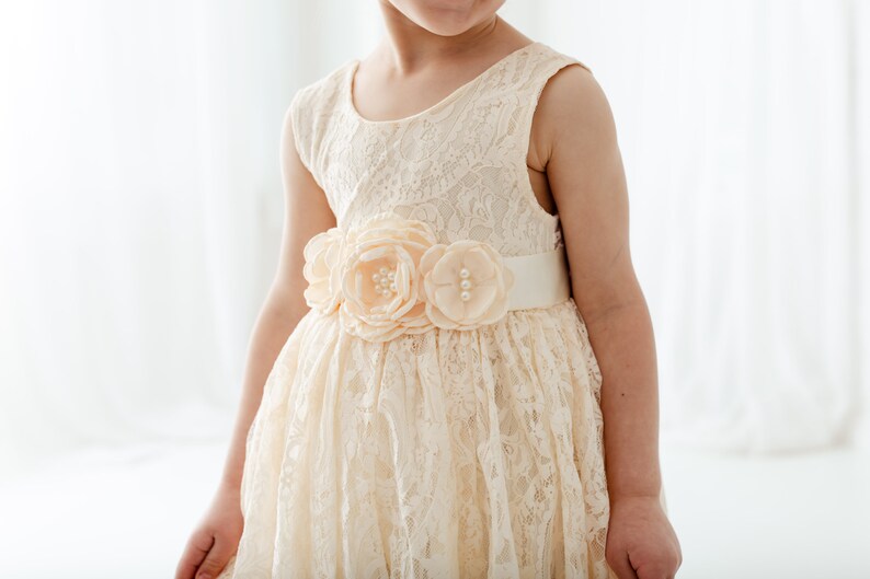 May include: A white lace dress with a peach-colored flower sash. The dress has a sleeveless design and a full skirt.