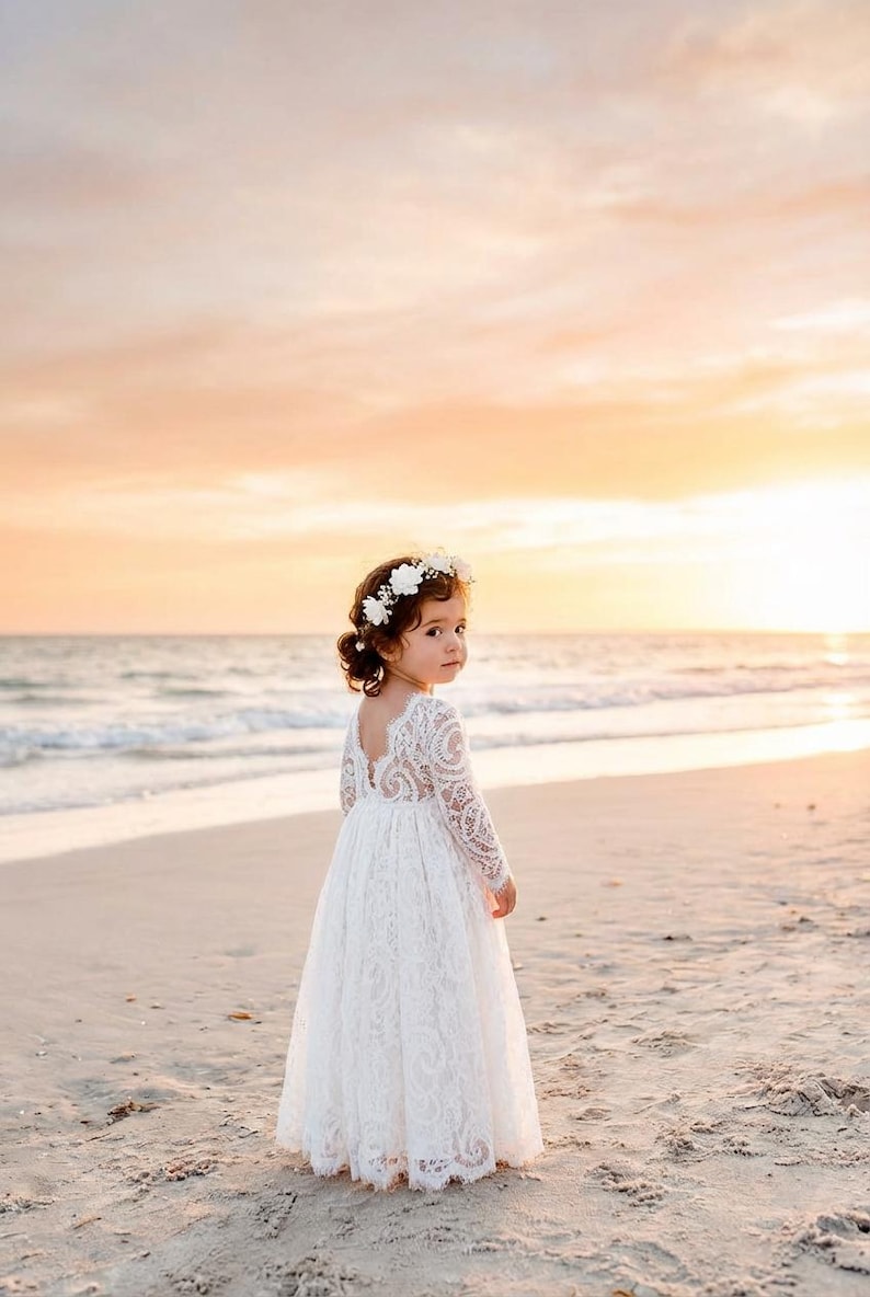 May include: A white long-sleeved dress featuring intricate lace detailing, a V-shaped back, and a full-length skirt. The garment is paired with a floral crown consisting of small white flowers worn on the head, completing the ensemble.