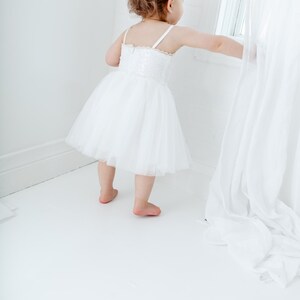 Puede incluir: Una ni&ntilde;a peque&ntilde;a con un vestido blanco de tul con detalles de encaje se encuentra en una habitaci&oacute;n blanca, mirando por una ventana. La ni&ntilde;a est&aacute; estirando la mano para tocar una cortina blanca.