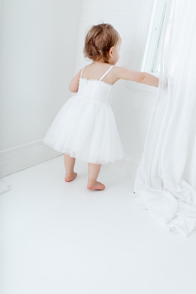 Puede incluir: Una ni&ntilde;a peque&ntilde;a con un vestido blanco de tul con detalles de encaje se encuentra en una habitaci&oacute;n blanca, mirando por una ventana. La ni&ntilde;a est&aacute; estirando la mano para tocar una cortina blanca.