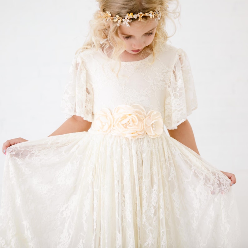 以下が含まれることがあります： 白いレースのドレスを着て、花柄のサッシュと金の冠をかぶった若い女の子