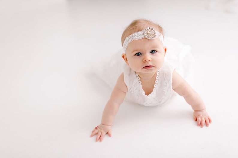 May include: A baby wearing a white dress with lace trim and a white headband with a jeweled embellishment. The baby is crawling on a white surface.