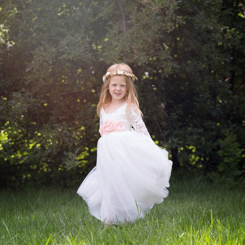 May include: A young girl wearing a white tulle dress with lace sleeves and a pink flower sash. She is wearing a flower crown and smiling at the camera.