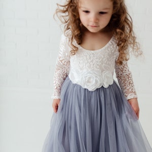 May include: A young girl wearing a white lace long-sleeved dress with a large white flower on the bodice and a grey tulle skirt.