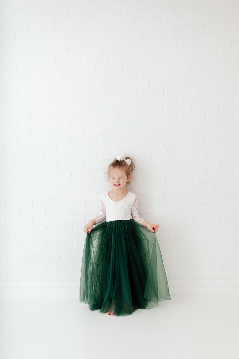 K&ouml;nnte beinhalten: Ein junges M&auml;dchen tr&auml;gt ein wei&szlig;es Spitzenoberteil mit langen &Auml;rmeln und einen gr&uuml;nen T&uuml;llrock. Sie steht vor einer wei&szlig;en Backsteinwand.