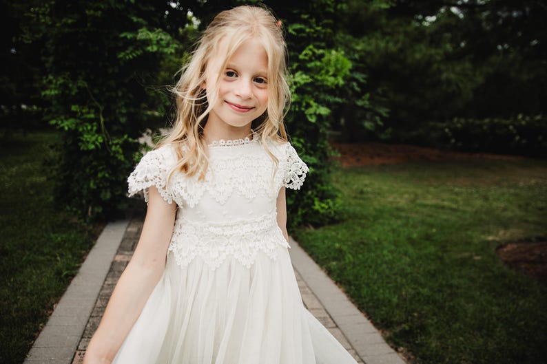 May include: A young girl with blonde hair smiles, wearing a white dress with lace detailing. The dress features a high neckline and short sleeves. She stands on a stone path, with green foliage in the background.