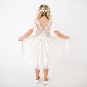 May include: An ivory-colored dress featuring a lace bodice with short sleeves and a deep V-shaped back. The skirt is made of layered lace with a scalloped hemline. The dress is paired with white shoes and a white ribbon tied in the hair.