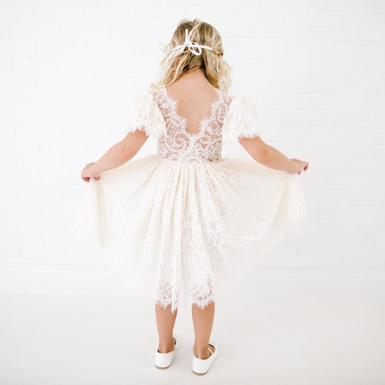 May include: An ivory-colored dress featuring a lace bodice with short sleeves and a deep V-shaped back. The skirt is made of layered lace with a scalloped hemline. The dress is paired with white shoes and a white ribbon tied in the hair.