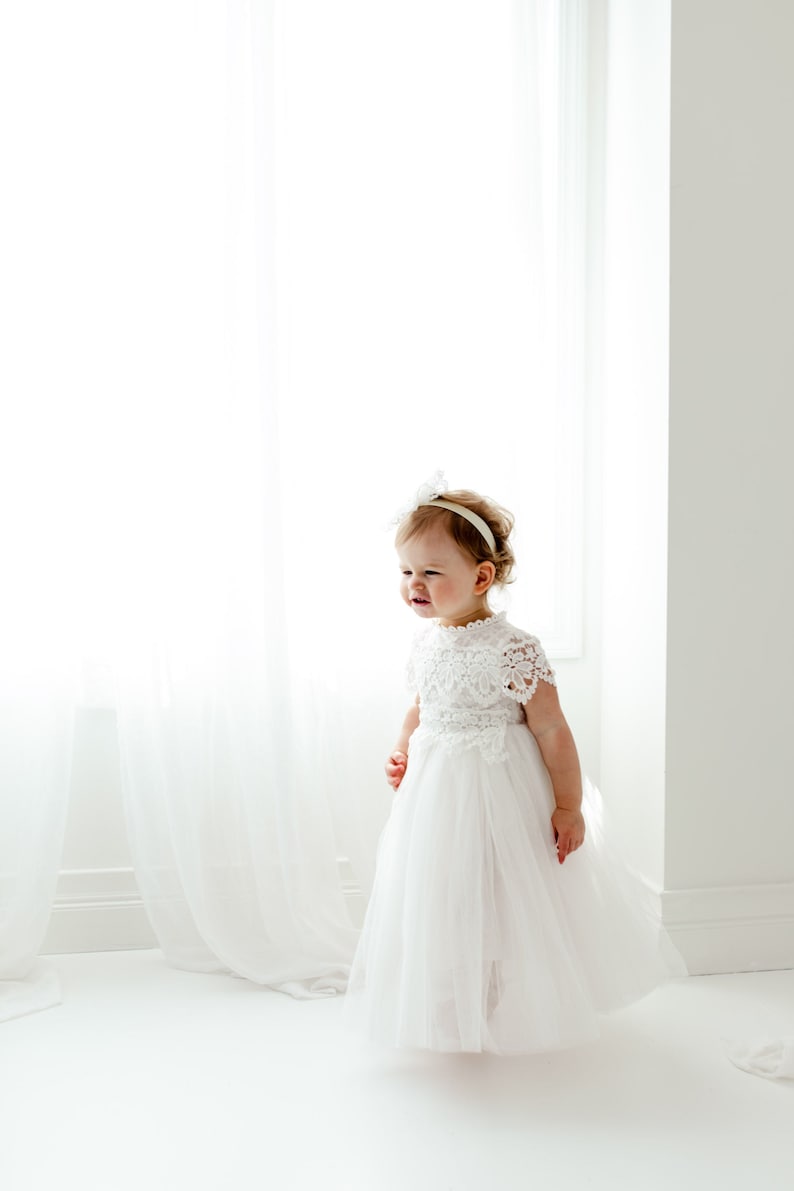 May include: A white lace and tulle dress with a matching headband. The dress has a detailed lace bodice and a flowing tulle skirt. The child is smiling, standing in front of a bright window.