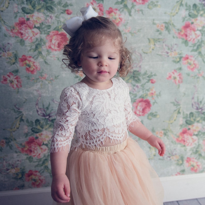 May include: A white lace top with three-quarter length sleeves and a scalloped hem paired with a peach-colored tulle skirt. A white hair bow is attached to the hair. The outfit features intricate floral lace patterns and layered tulle fabric.