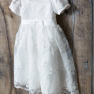 May include: A white dress with short puff sleeves, a round neckline, and a satin waistband featuring a small center bow. The bodice and skirt are adorned with intricate white floral embroidery and lace overlays, hanging on a bright pink hanger.