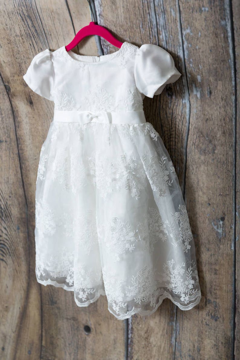 May include: A white dress with short puff sleeves, a round neckline, and a satin waistband featuring a small center bow. The bodice and skirt are adorned with intricate white floral embroidery and lace overlays, hanging on a bright pink hanger.