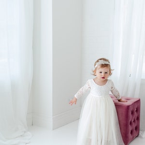 May include: A white long-sleeved dress with a lace bodice and a layered tulle skirt. The dress features a scoop neckline and a fitted waist. A decorative white headband with intricate floral lace detailing is worn on the head.