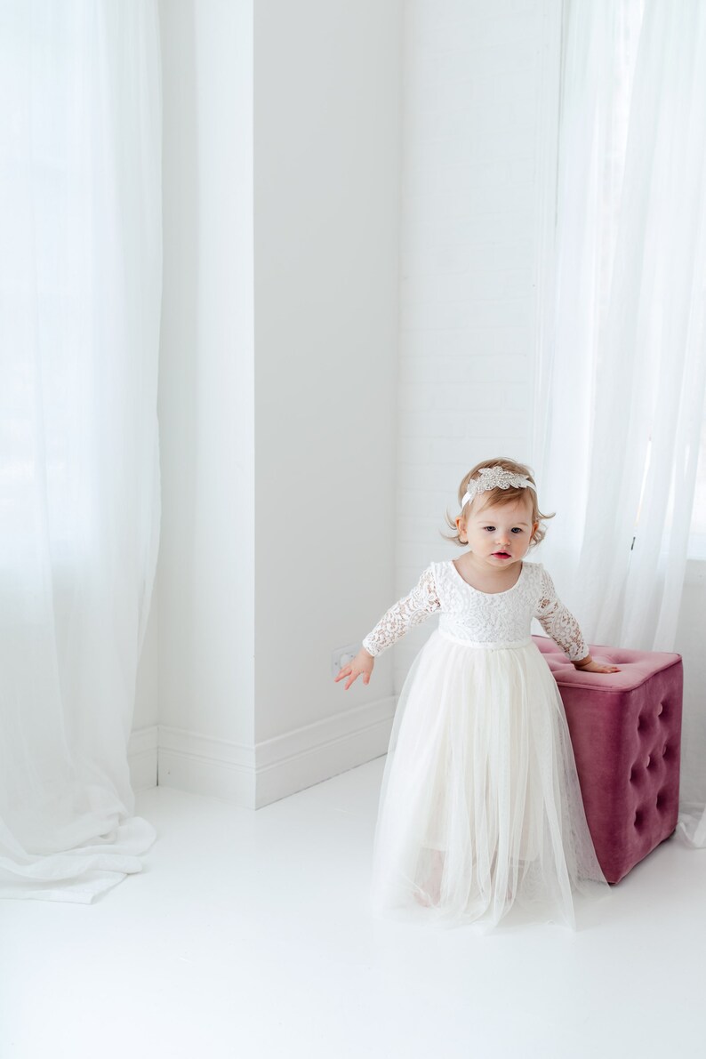 May include: A white long-sleeved dress with a lace bodice and a layered tulle skirt. The dress features a scoop neckline and a fitted waist. A decorative white headband with intricate floral lace detailing is worn on the head.