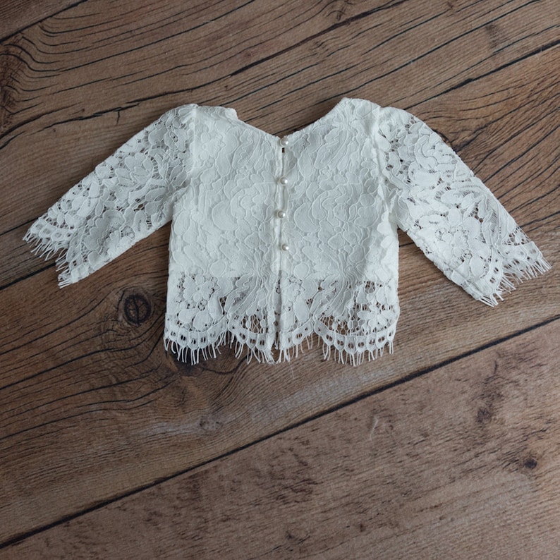 May include: A white long-sleeved top crafted from intricate floral lace fabric. The garment features a scalloped hemline with fringe detailing and a central vertical placket on the front secured by four small, round, white pearl-like buttons.