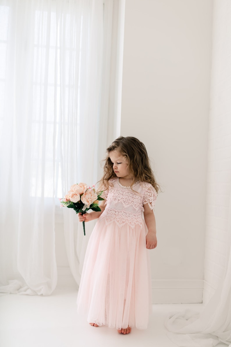 May include: A pale pink dress featuring a lace bodice with short sleeves and a round neckline adorned with small pearls. The skirt is made of layered tulle, falling to the floor. The dress is paired with a small bouquet of artificial pink flowers.