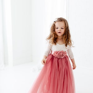 May include: A young girl wearing a white lace top with a pink tulle skirt and a large pink flower accent at the waist.