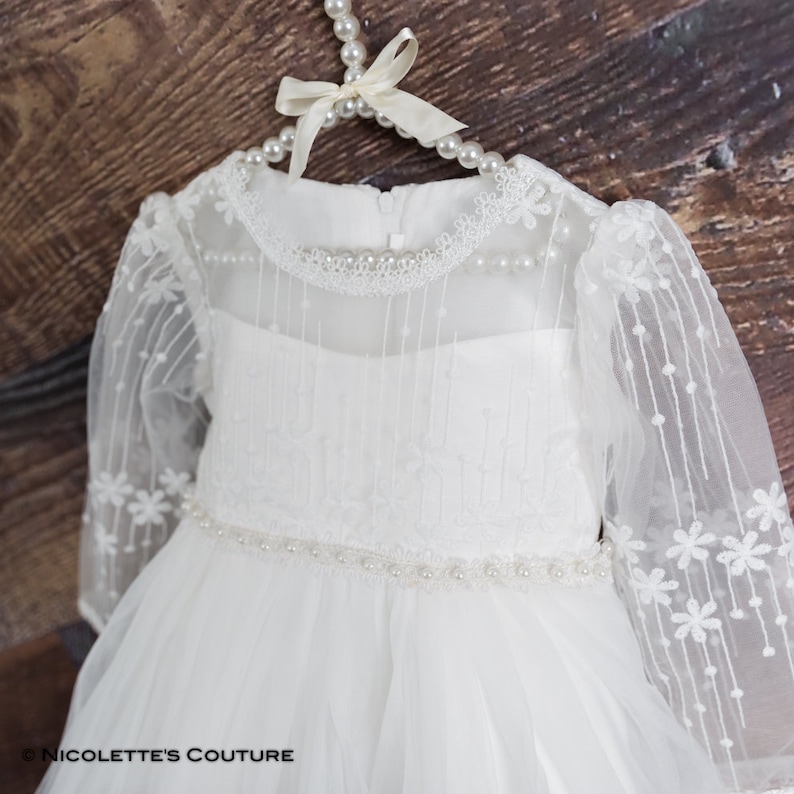 K&ouml;nnte beinhalten: Wei&szlig;es T&uuml;ll-Babykleid mit langen &Auml;rmeln, Perlenbesatz und einer floralen Spitzen&uuml;berlage. Das Kleid befindet sich auf einem Kleiderb&uuml;gel mit Perlen- und Schleifendetail.
