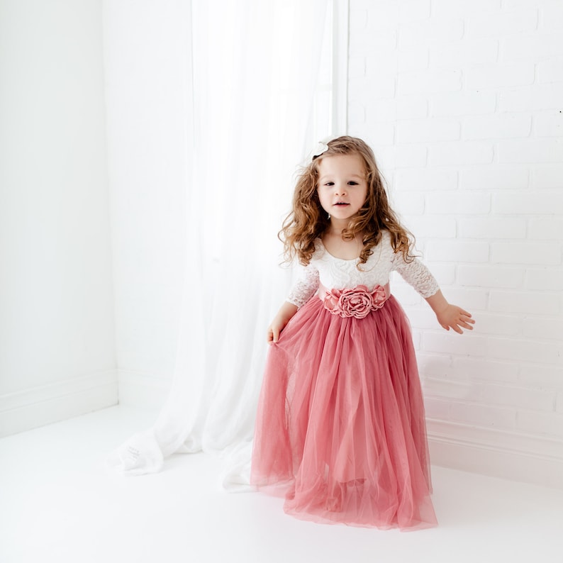 May include: A young girl wearing a white lace top with a pink tulle skirt and a large pink flower on the waist.