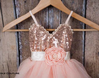 May include: A peach-colored dress with a sequined bodice and a tulle skirt, hanging on a wooden hanger. The bodice is adorned with rose gold sequins and a white ribbon belt. A large, peach-colored flower with a pearl center accents the waist.