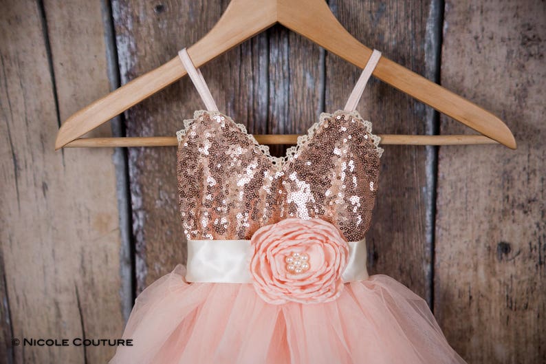 May include: A blush pink tulle dress with a rose gold sequin bodice and a large blush pink flower accent. The dress is on a wooden hanger.