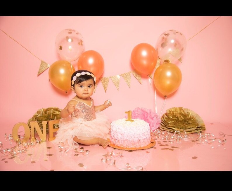 May include: A baby girl wearing a pink sequined dress and a flower crown sits in front of a pink cake decorated with a gold number one. The cake is surrounded by gold and pink balloons and confetti.