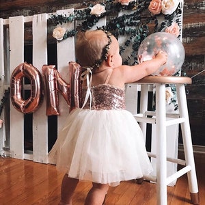 May include: A young child wearing a white tulle dress with a rose gold sequin bodice and a rose gold crown. The child is standing in front of a white pallet wall with rose gold foil balloons that spell out "ONE".