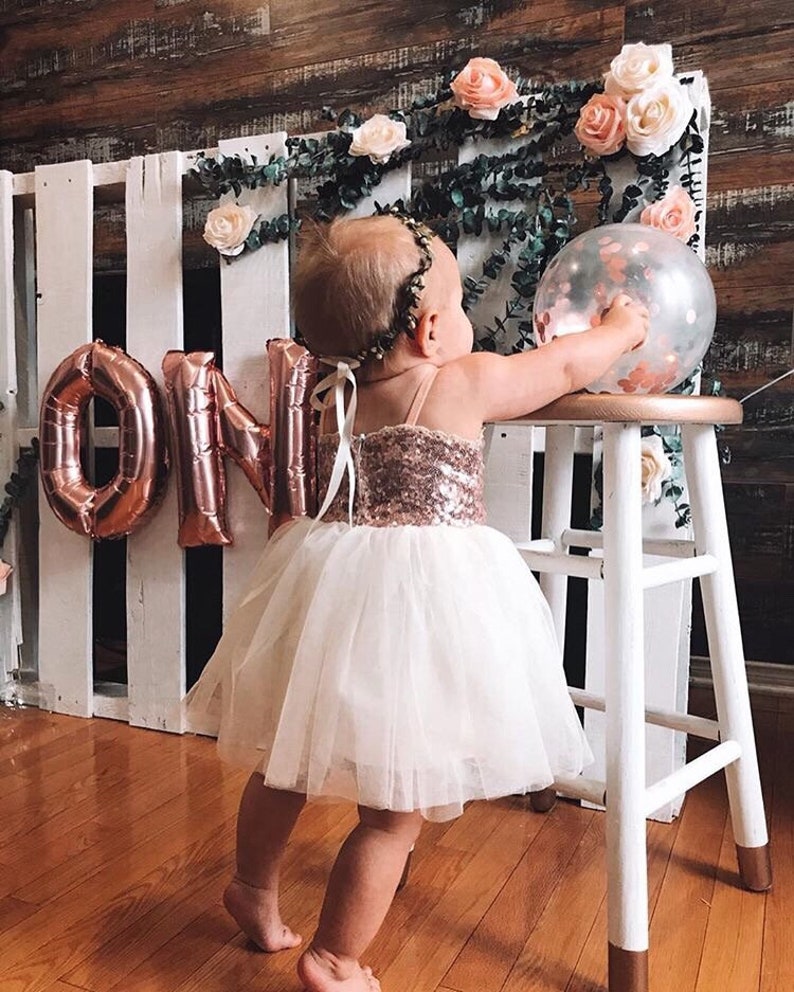 May include: A young child wearing a white tulle dress with a rose gold sequin bodice and a rose gold crown. The child is standing in front of a white pallet wall with rose gold foil balloons that spell out "ONE".