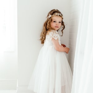 May include: A young girl wearing a white tulle dress with lace detailing and a gold flower crown. She is standing in a white room with a white curtain behind her.