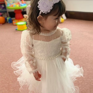 May include: A white dress featuring sheer puff sleeves with floral embroidery, a bodice with vertical pin-tuck detailing, and a layered tulle skirt accented by a pearl-beaded waistline. A matching white headband with two fabric roses and a central rhinestone embellishment completes the ensemble.