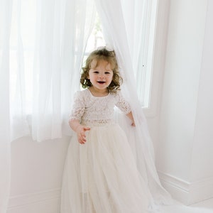 May include: A young child wearing a cream-colored lace bodice dress with a tulle skirt, standing near a window with white curtains. The dress has short sleeves and a square neckline. The child is smiling.