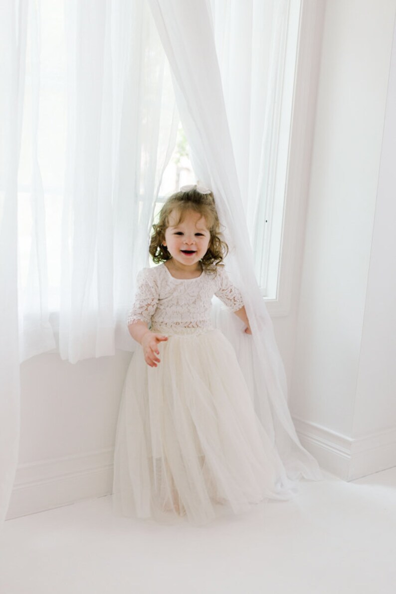 May include: A young child wearing a cream-colored lace bodice dress with a tulle skirt, standing near a window with white curtains. The dress has short sleeves and a square neckline. The child is smiling.