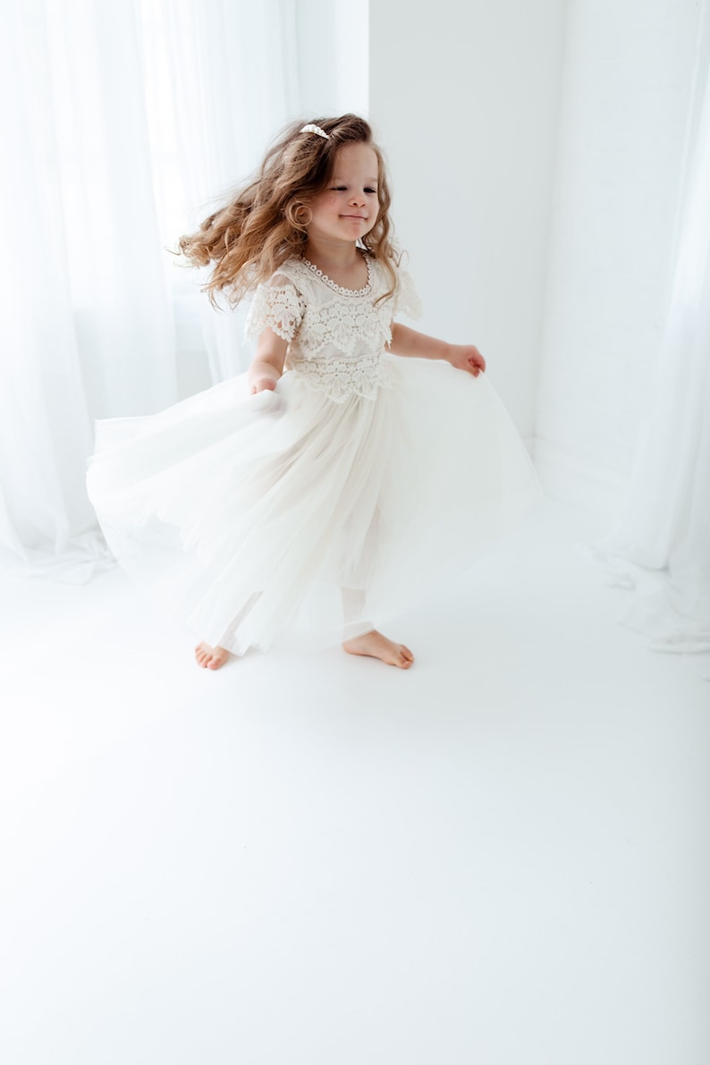 Op de afbeelding: Een jong meisje met lang krullend haar draagt een witte tule jurk met kantdetails. Ze draait rond en glimlacht in een witte kamer.