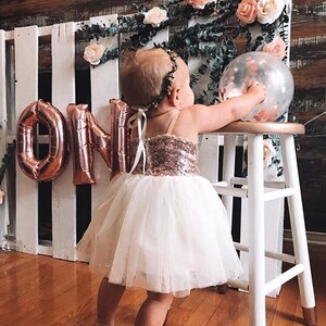 May include: A young child wearing a white tulle dress with a rose gold sequin bodice and a rose gold crown. The child is standing in front of a white pallet wall with rose gold foil balloons spelling out "ONE".